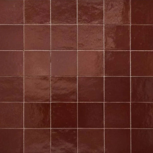 Glossy burgundy porcelain tiles inspired by Moroccan zellige, featuring subtly uneven edges and rich tonal variation. The tiles reflect light across a textured surface, creating a handcrafted, artisanal look when used on walls in a decorative interior setting.