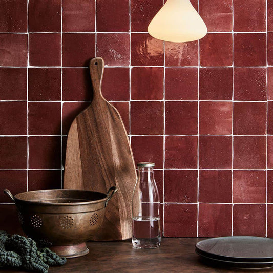 Glossy burgundy porcelain tiles inspired by Moroccan zellige, featuring subtly uneven edges and rich tonal variation. The tiles reflect light across a textured surface, creating a handcrafted, artisanal look when used on walls in a decorative interior setting.
